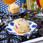 Scones on blue and white plates with glasses of juice on a metal table outdoors.