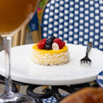 Small fruit tart on a white plate with a glass of orange juice and a checkered chair in the background.