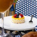 Small fruit tart on a white plate with a glass of orange juice and a checkered chair in the background.