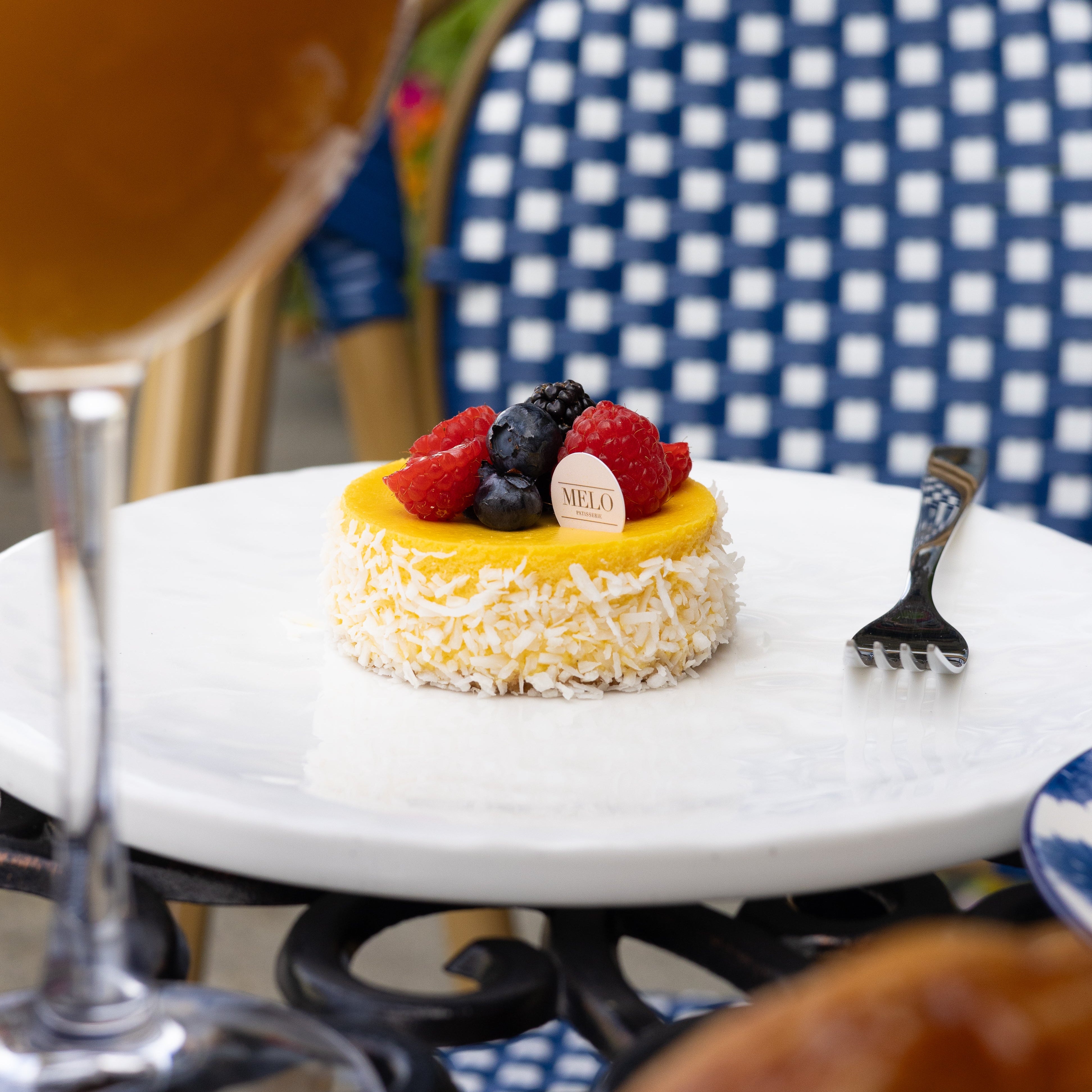 Small fruit tart on a white plate with a glass of orange juice and a checkered chair in the background.