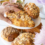 Hand reaching for a croissant on a white plate with other pastries, flowers, and a cup in the background.