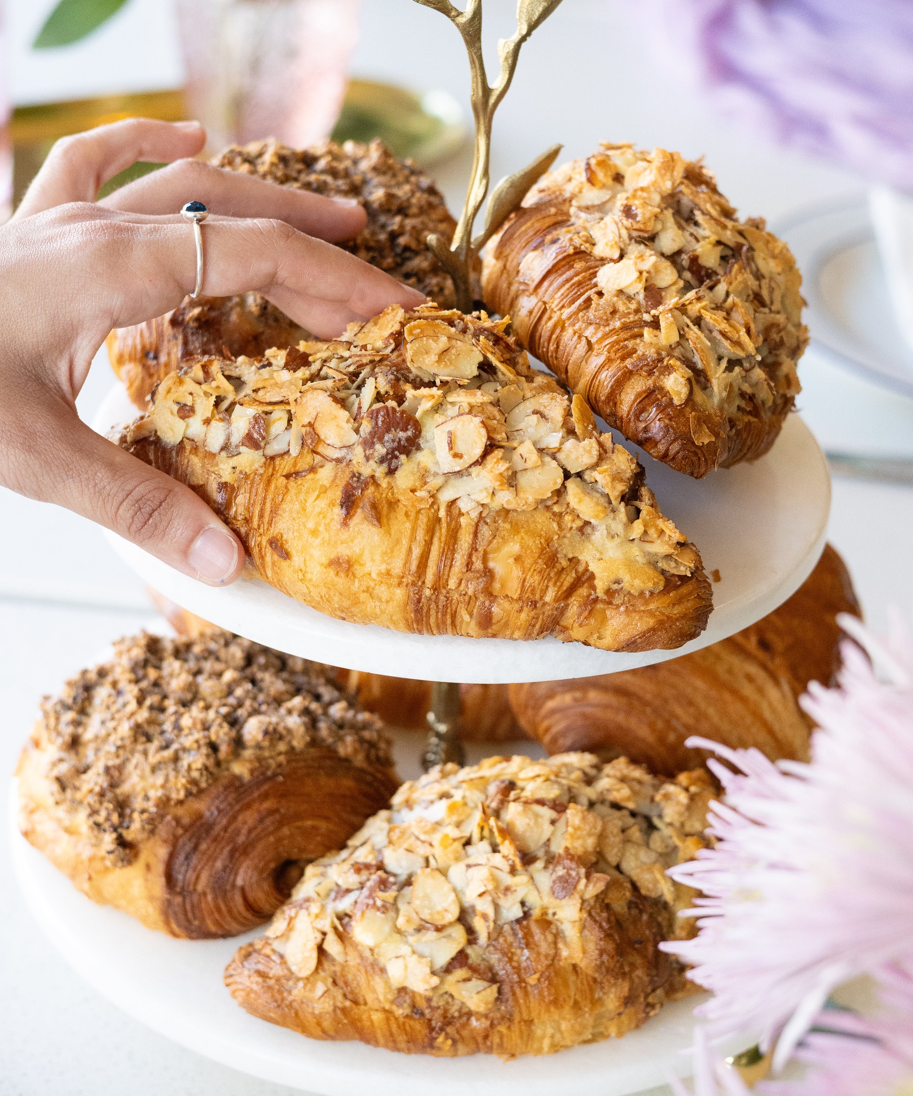 Hand reaching for a croissant on a white plate with other pastries, flowers, and a cup in the background.