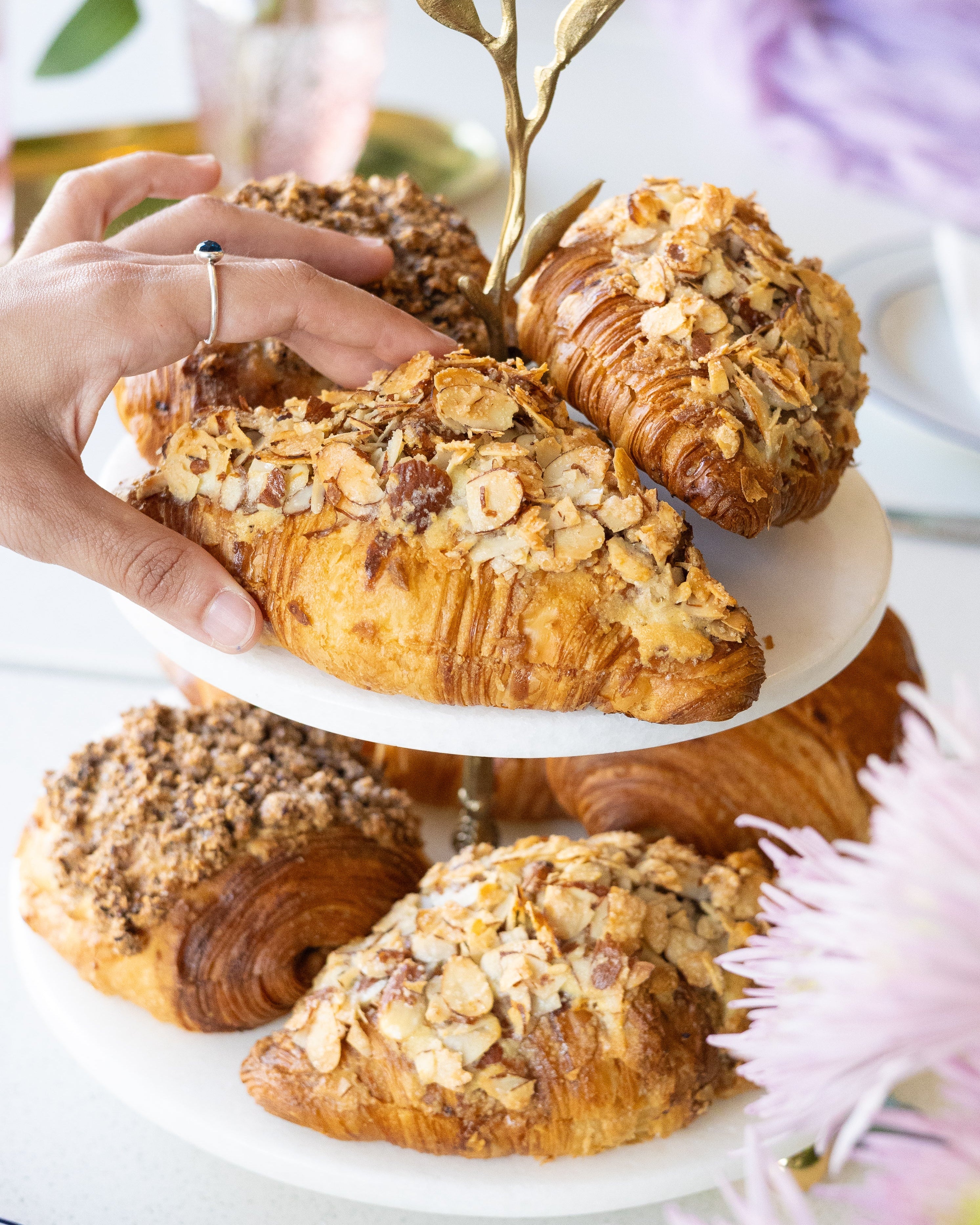 Hand reaching for a croissant on a white plate with other pastries, flowers, and a cup in the background.