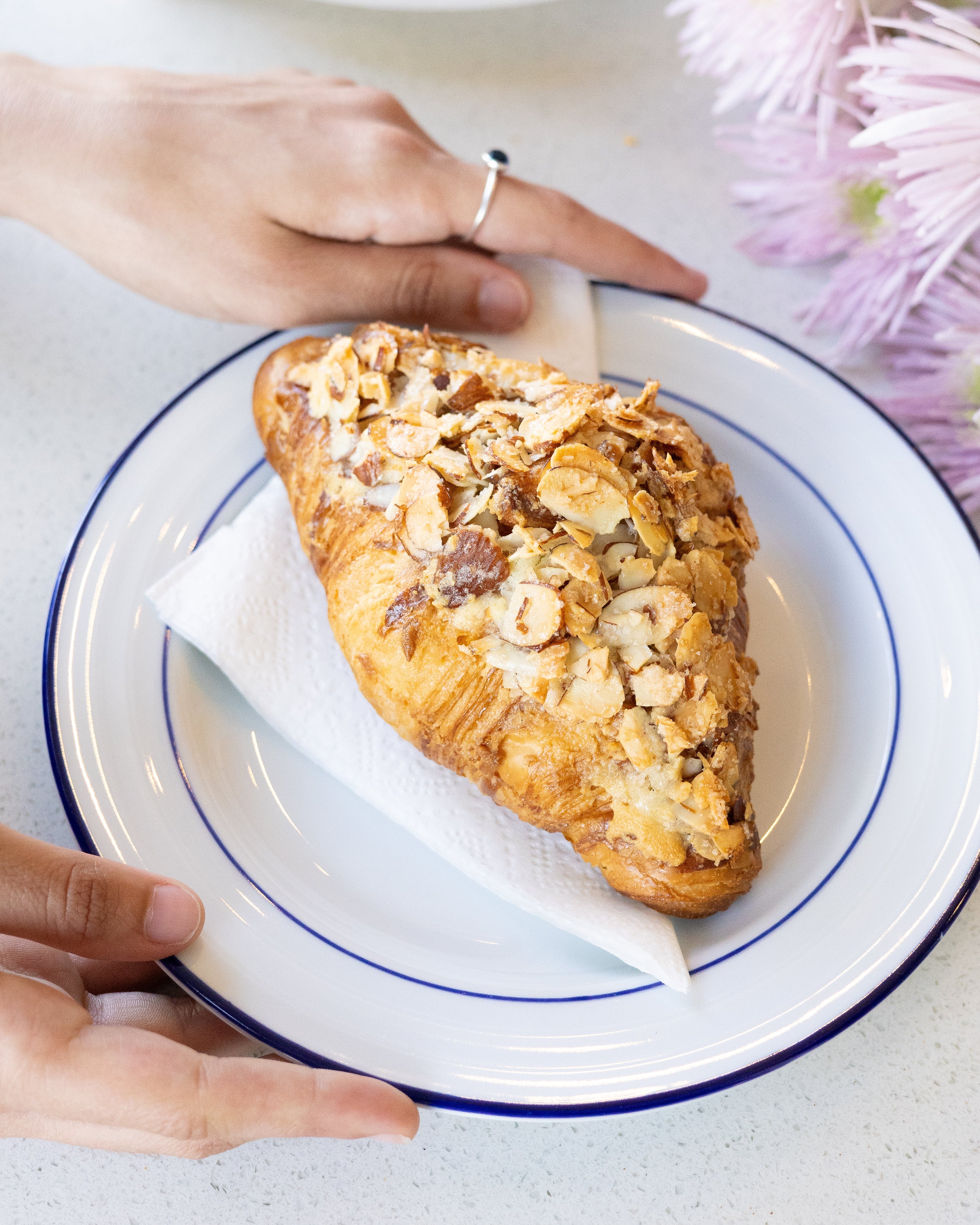 Pastry on a plate with hands holding it, surrounded by flowers and more pastries.