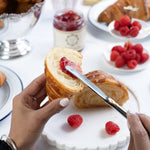 Person cutting a croissant with raspberry jam on a white plate with raspberries.