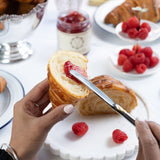 Person cutting a croissant with raspberry jam on a white plate with raspberries.