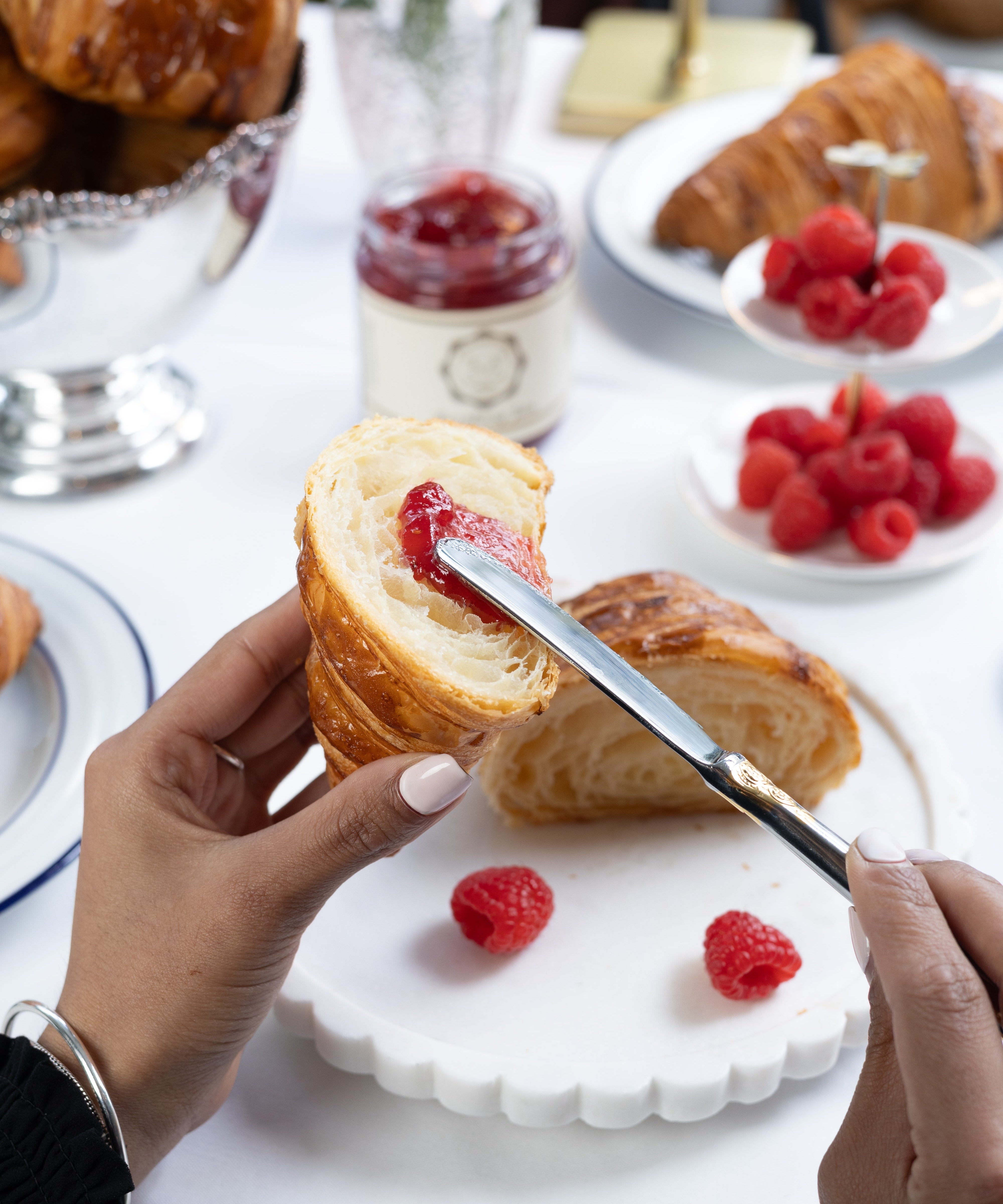 Person cutting a croissant with raspberry jam on a white plate with raspberries.
