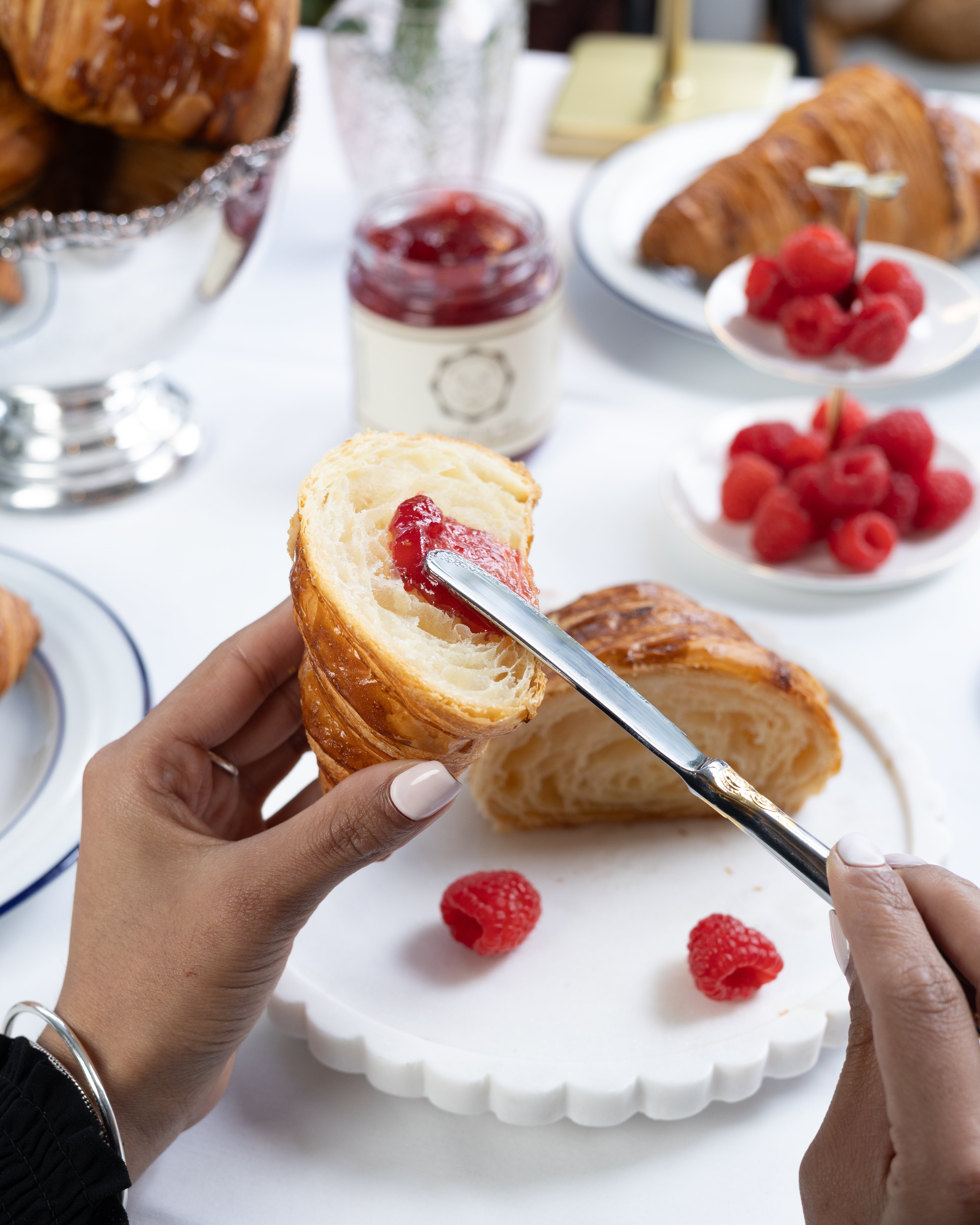 Person cutting a croissant with raspberry jam on a white plate with raspberries.