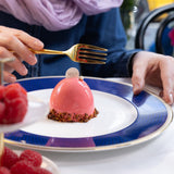 Person holding a gold fork over a pink dessert on a blue and white plate with raspberries.