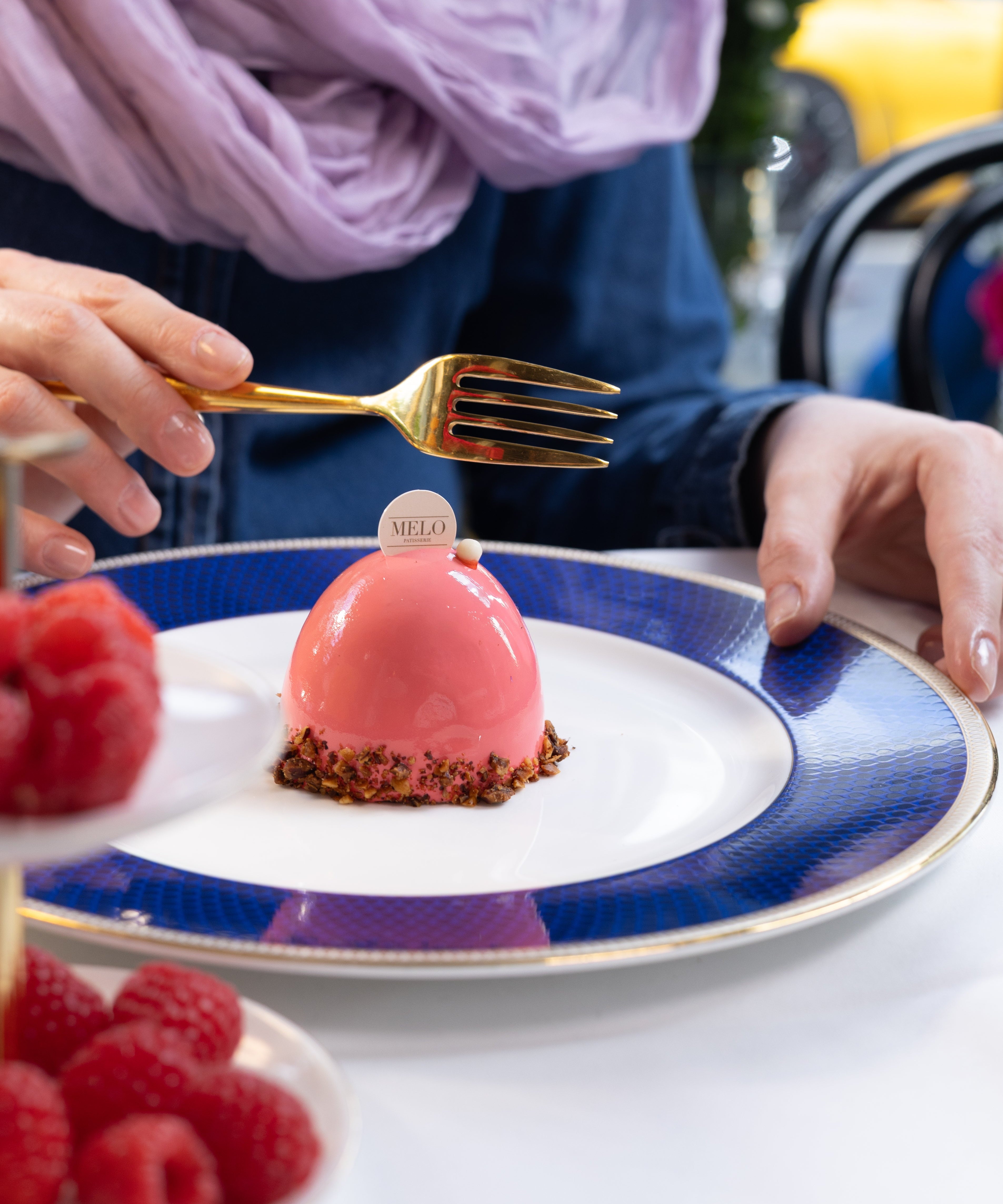 Person holding a gold fork over a pink dessert on a blue and white plate with raspberries.