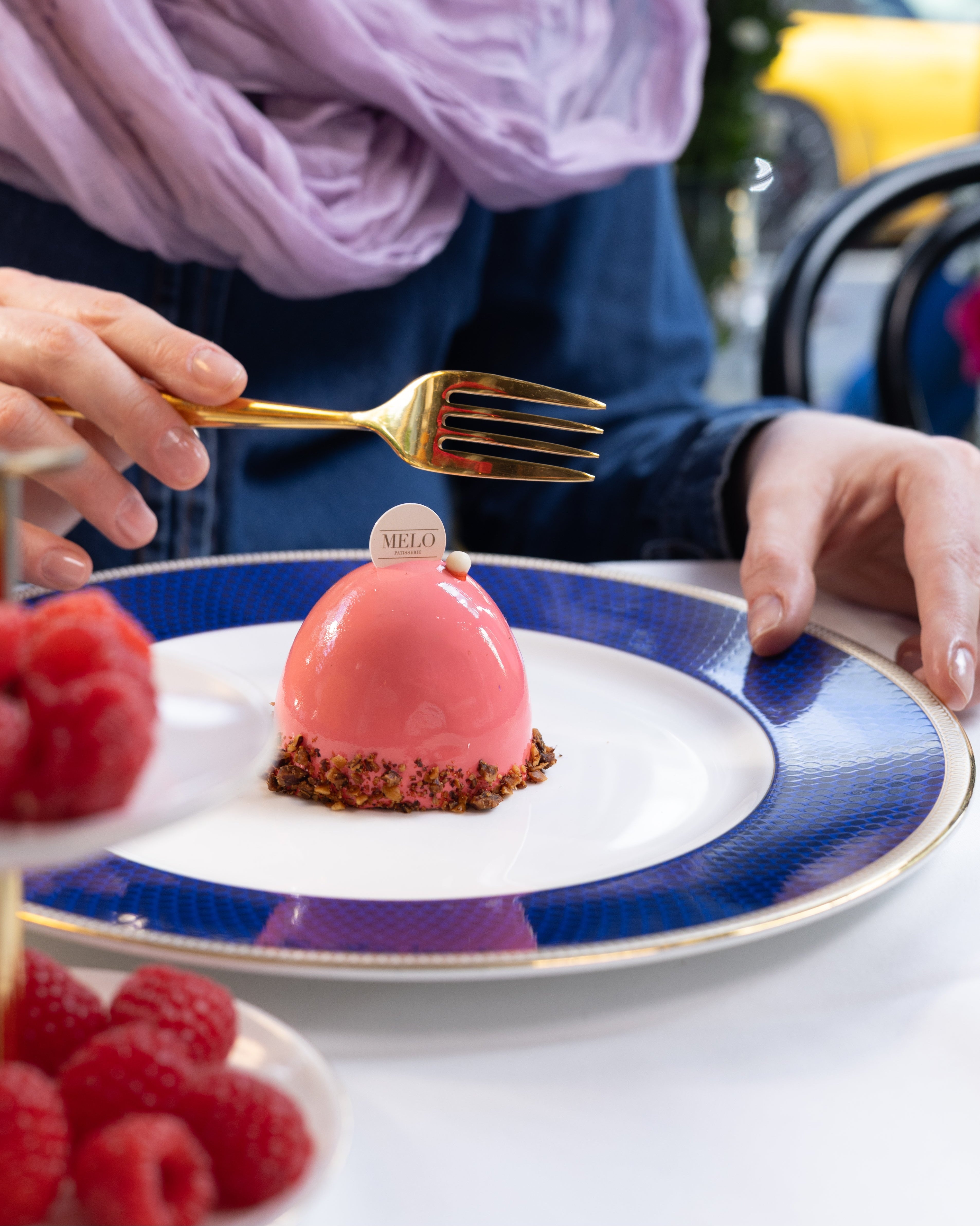 Person holding a gold fork over a pink dessert on a blue and white plate with raspberries.