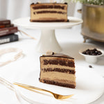 Slice of layered chocolate cake on a white plate with a gold fork, placed on a white surface.
