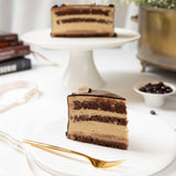 Slice of layered chocolate cake on a white plate with a gold fork, placed on a white surface.