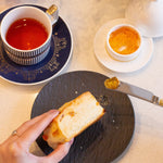 Tea cup with saucer, bread, and small dish on a table with a teapot and flowers.