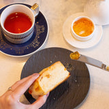 Tea cup with saucer, bread, and small dish on a table with a teapot and flowers.