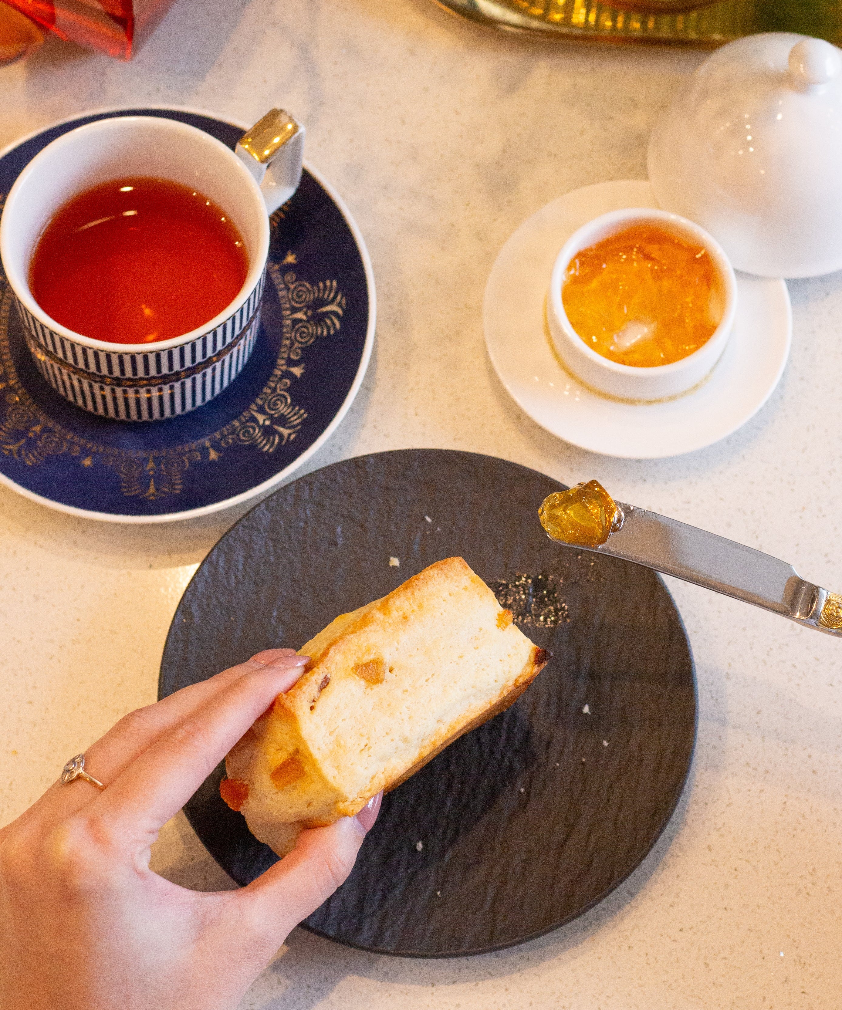 Tea cup with saucer, bread, and small dish on a table with a teapot and flowers.