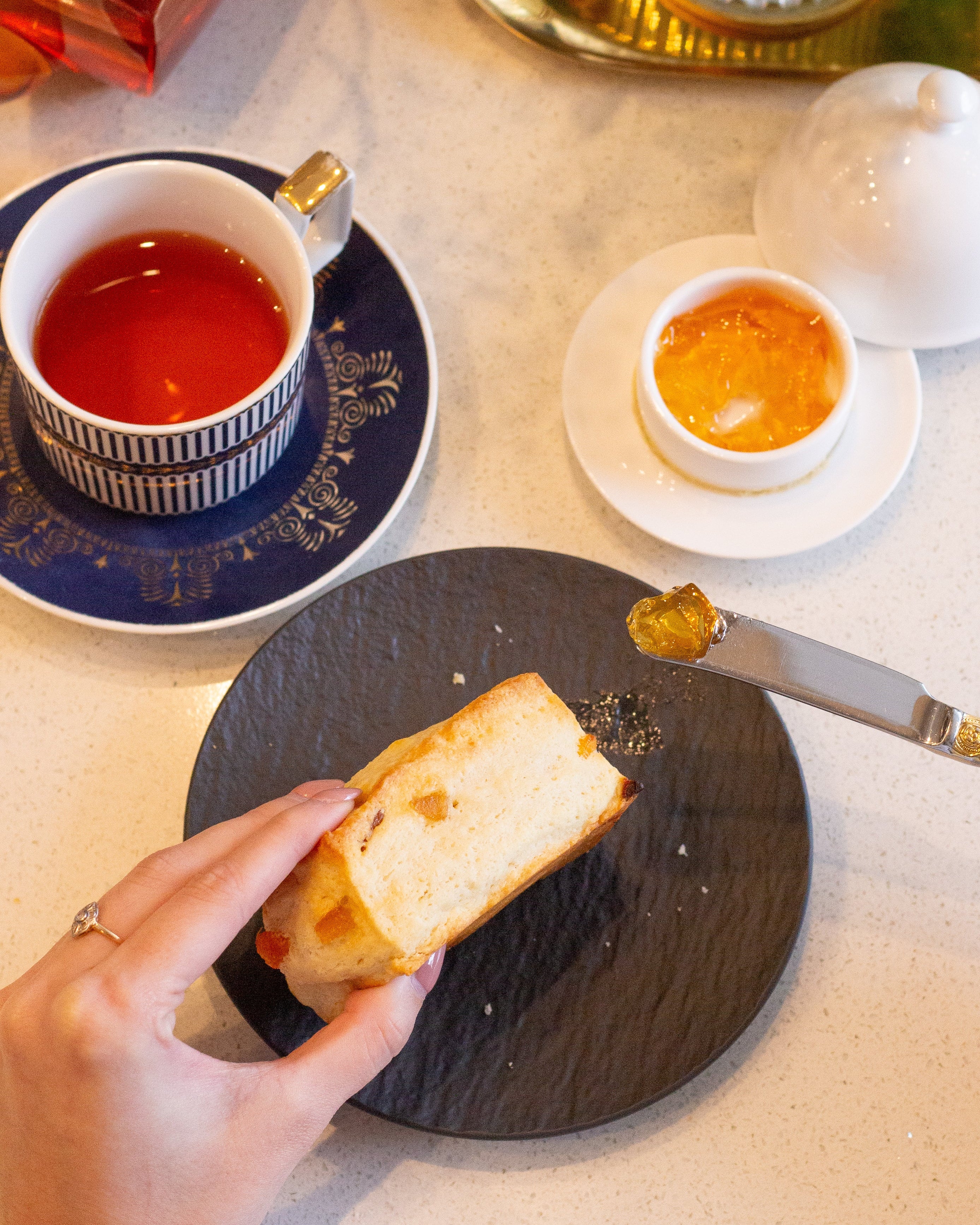 Tea cup with saucer, bread, and small dish on a table with a teapot and flowers.
