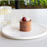 Small dessert with a cherry on top on a white plate, set against a blurred background of a table with a glass of water and a cup.