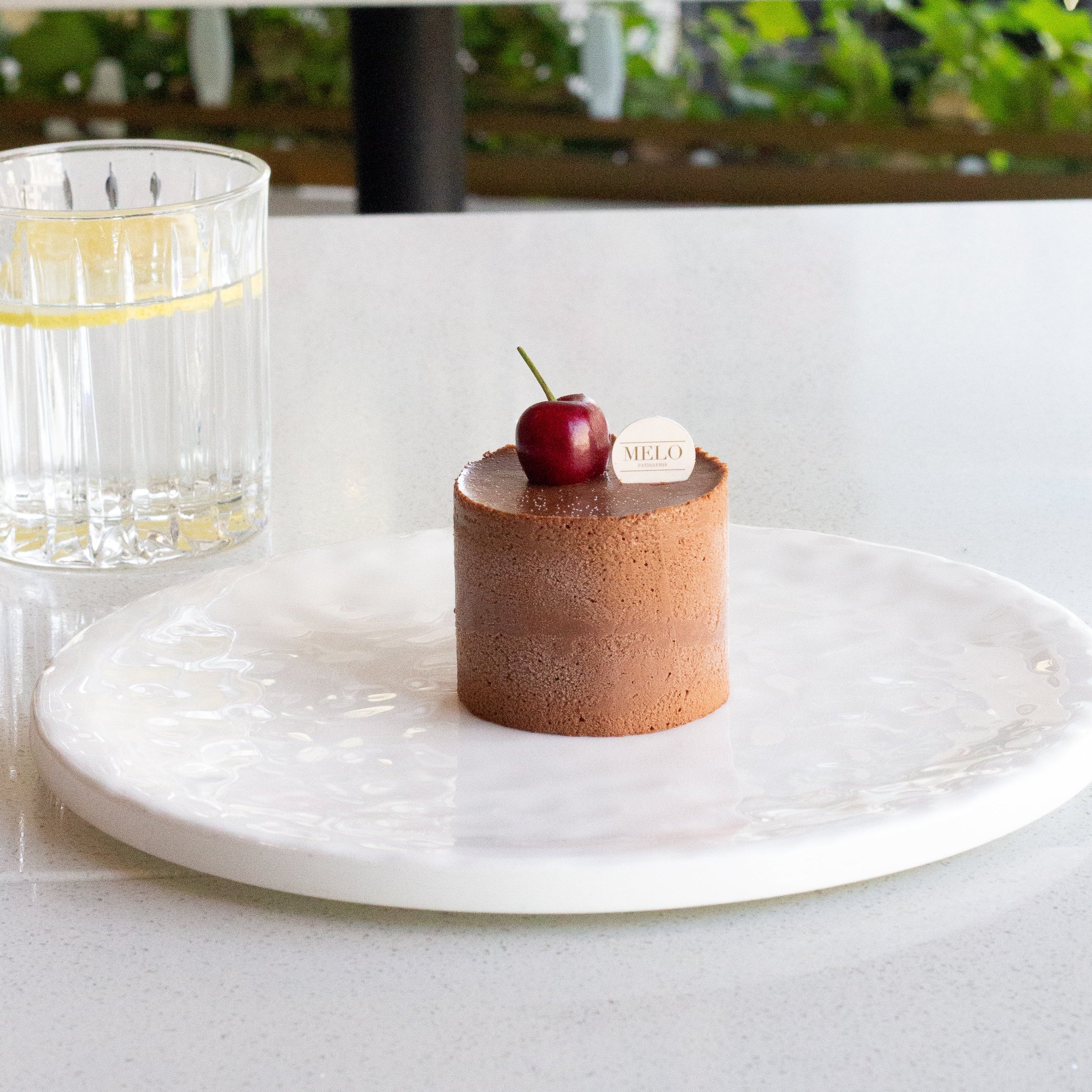 Small dessert with a cherry on top on a white plate, set against a blurred background of a table with a glass of water and a cup.