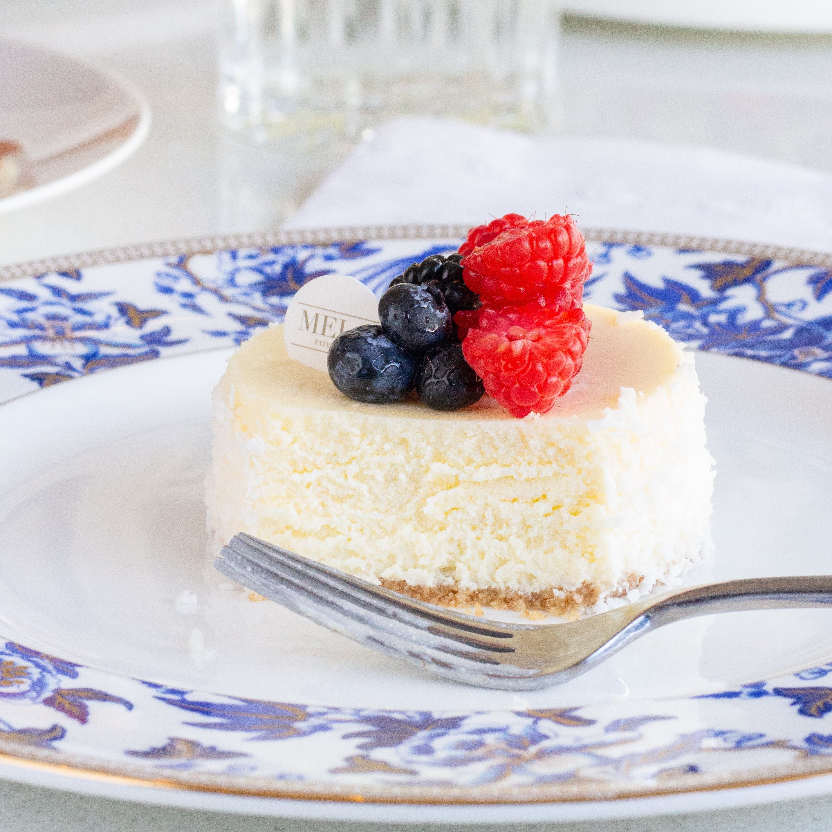 Small cheesecake with berries on a decorative plate with a fork, set against a blurred background of fruits and glasses.