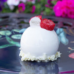 White dessert with raspberries and a 'Melo' label on a reflective surface with flowers in the background