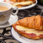 Croissant sandwich with cheese and ham on a plate with a cup of coffee in the background.