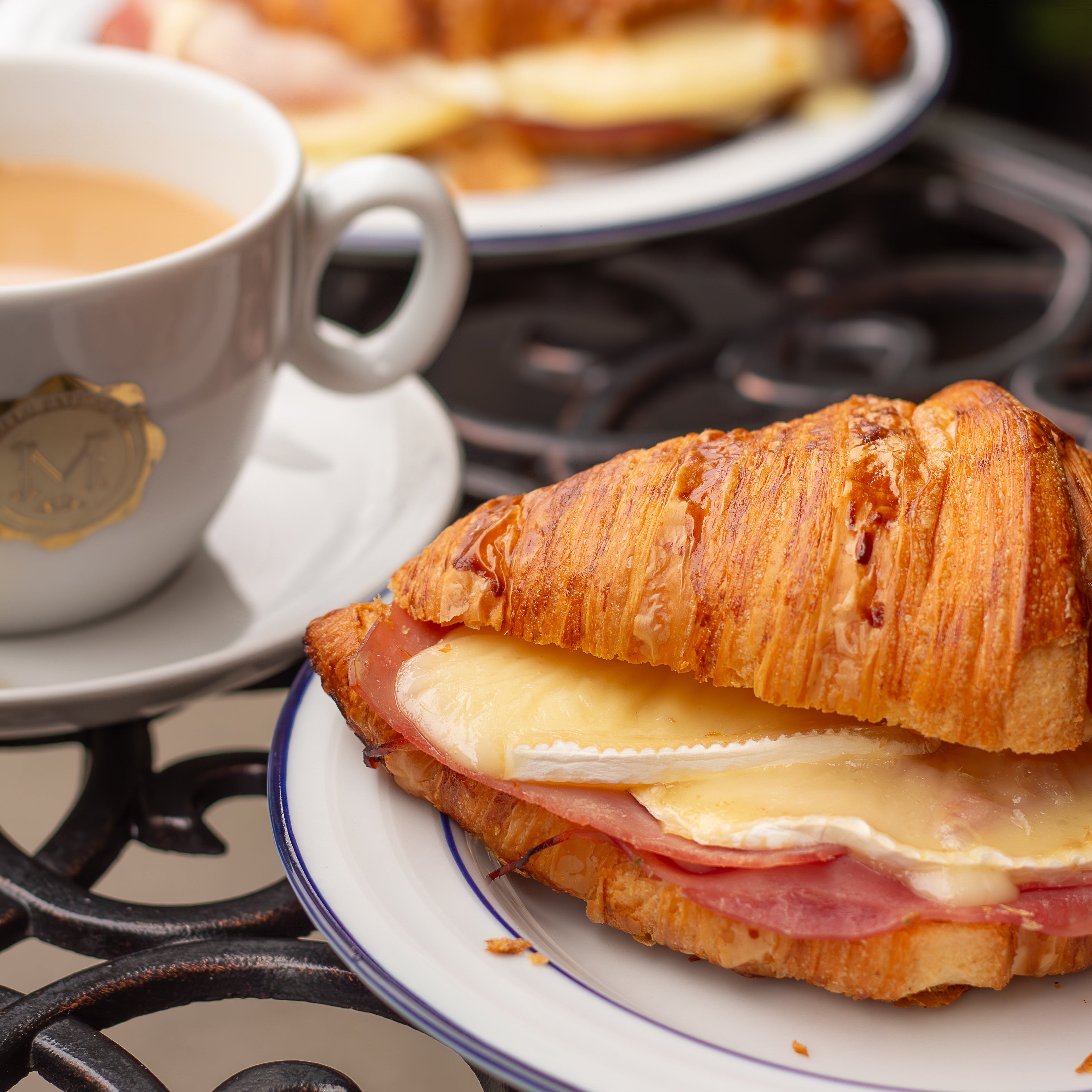 Croissant sandwich with cheese and ham on a plate with a cup of coffee in the background.