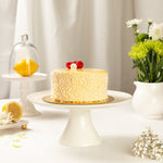 Yellow lemon cake on a white cake stand with lemons and flowers in the background