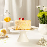 Yellow lemon cake on a white cake stand with lemons and flowers in the background