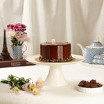 Chocolate cake on a white stand with flowers and teapots in the background