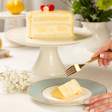 Slice of lemon cake on a decorative plate with a hand holding a gold fork, surrounded by flowers and a lemon.