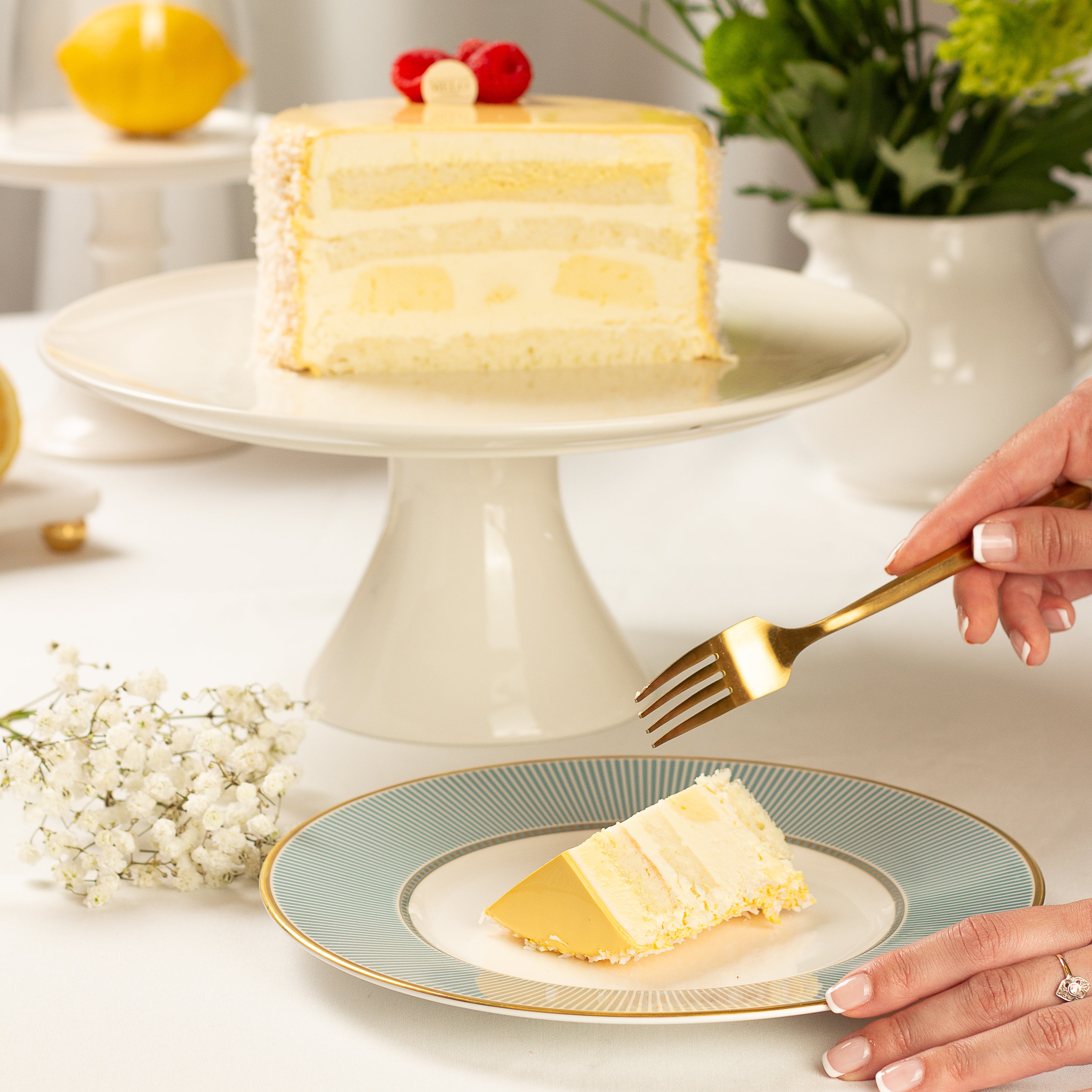 Slice of lemon cake on a decorative plate with a hand holding a gold fork, surrounded by flowers and a lemon.
