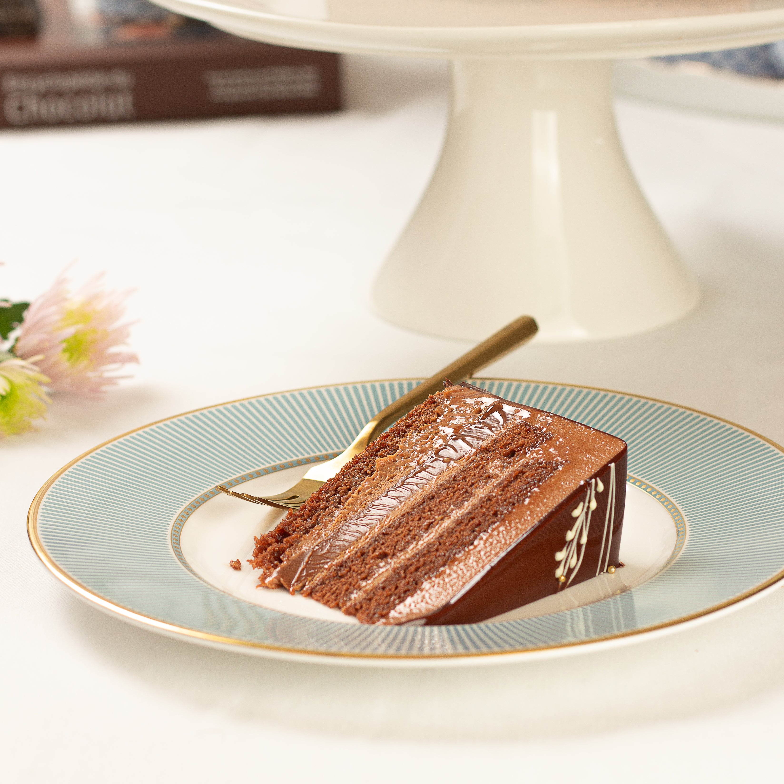 Slice of chocolate cake on a decorative plate with a cake stand in the background
