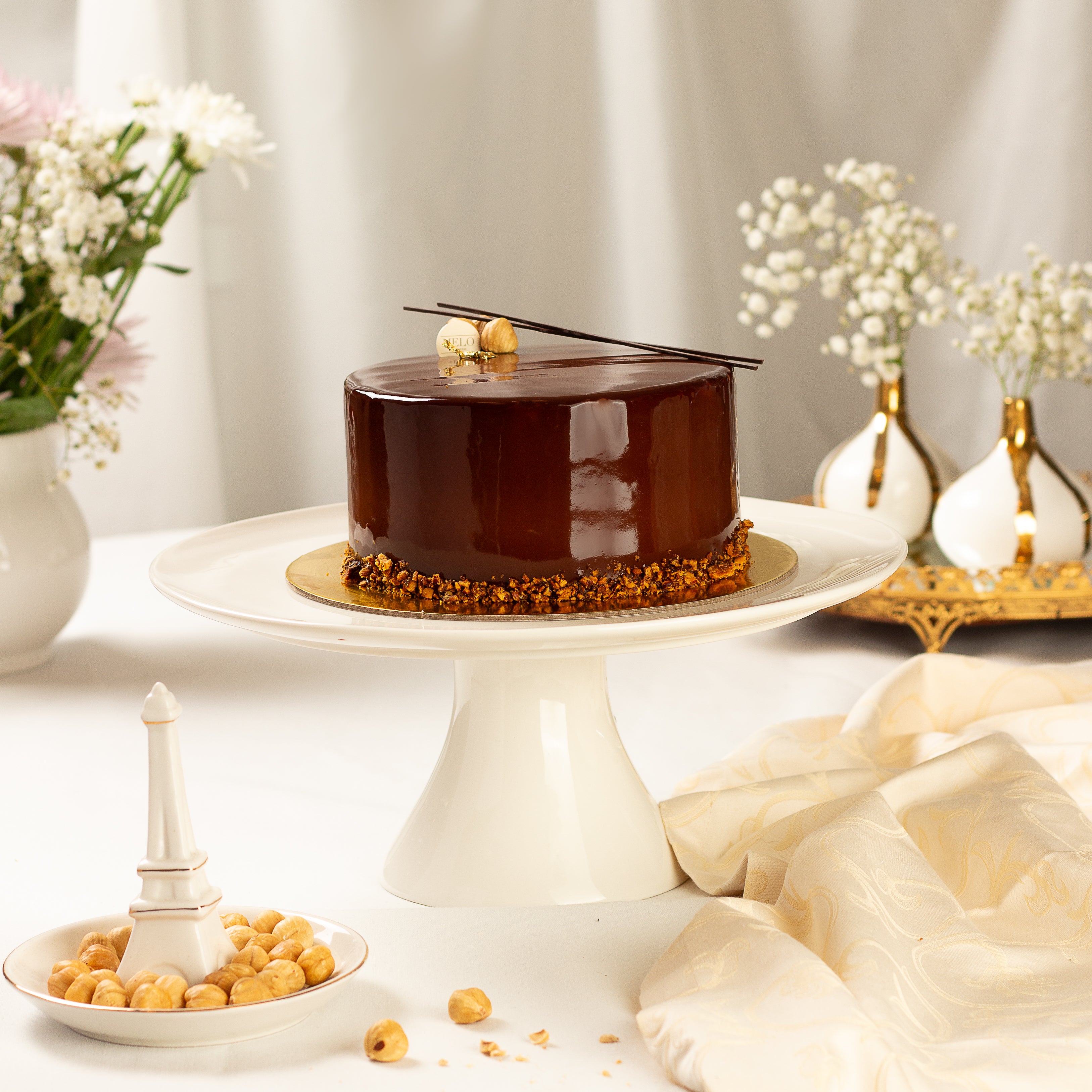 Chocolate cake on a white stand with decorative elements and flowers in the background