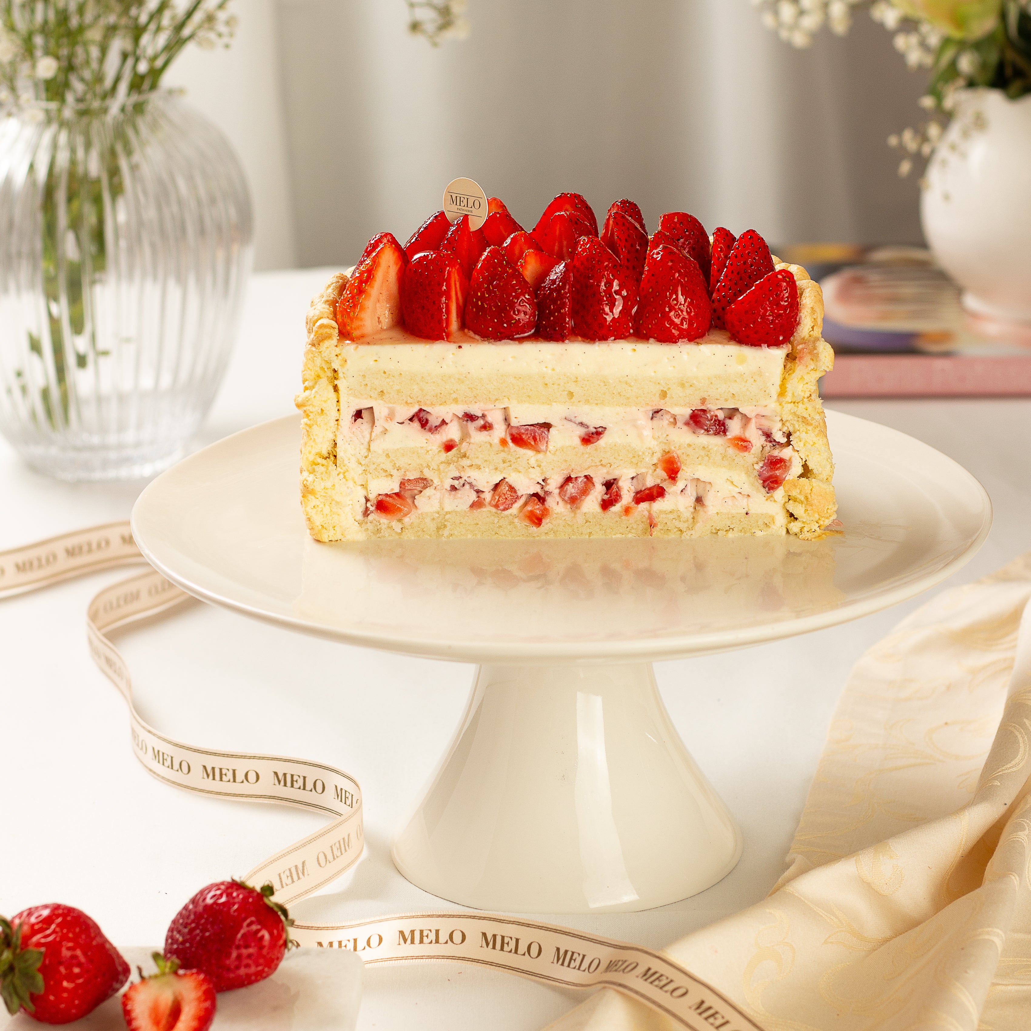 Strawberry cake on a white cake stand with decorative ribbons and flowers in the background.