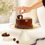 Chocolate cake on a white stand with a hand placing cherries on top, surrounded by flowers and a ribbon.