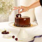 Chocolate cake on a white stand with a hand placing cherries on top, surrounded by flowers and a ribbon.