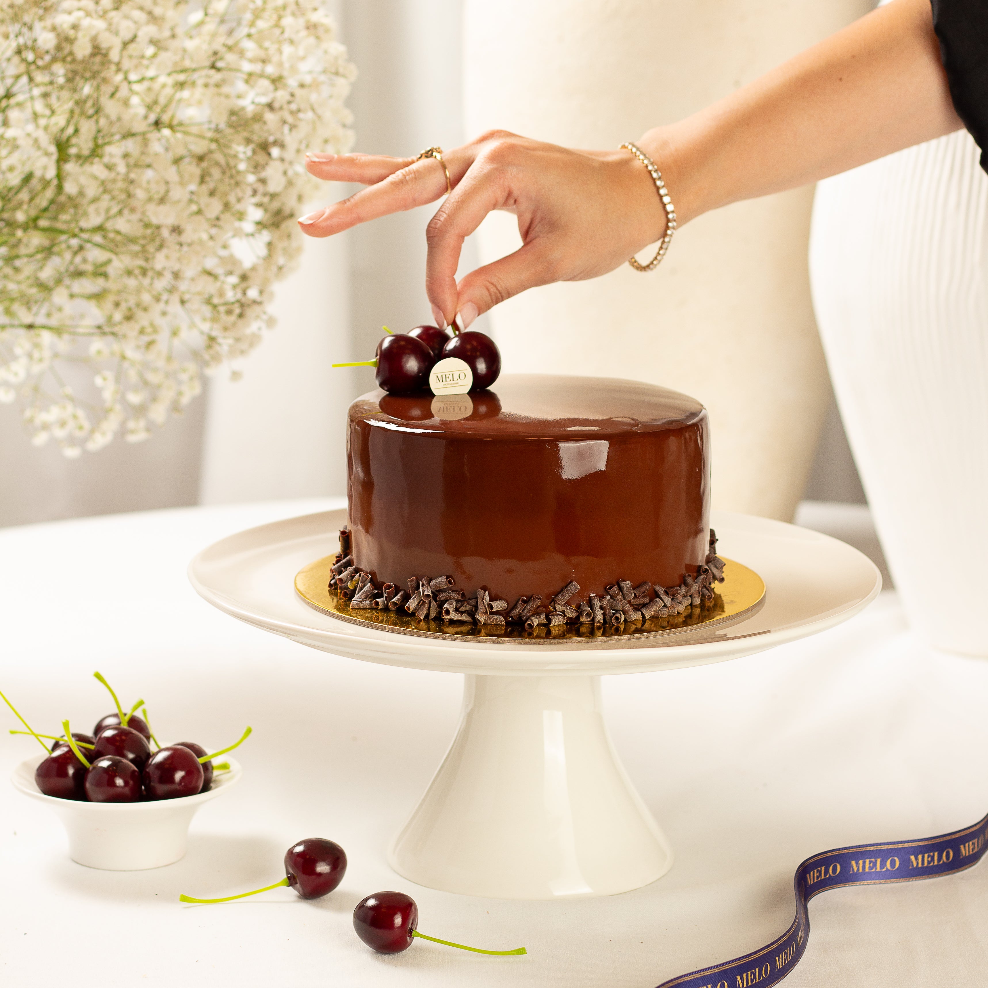 Chocolate cake on a white stand with a hand placing cherries on top, surrounded by flowers and a ribbon.