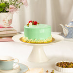 Green cake on a white cake stand with a teacup and tea set in the background