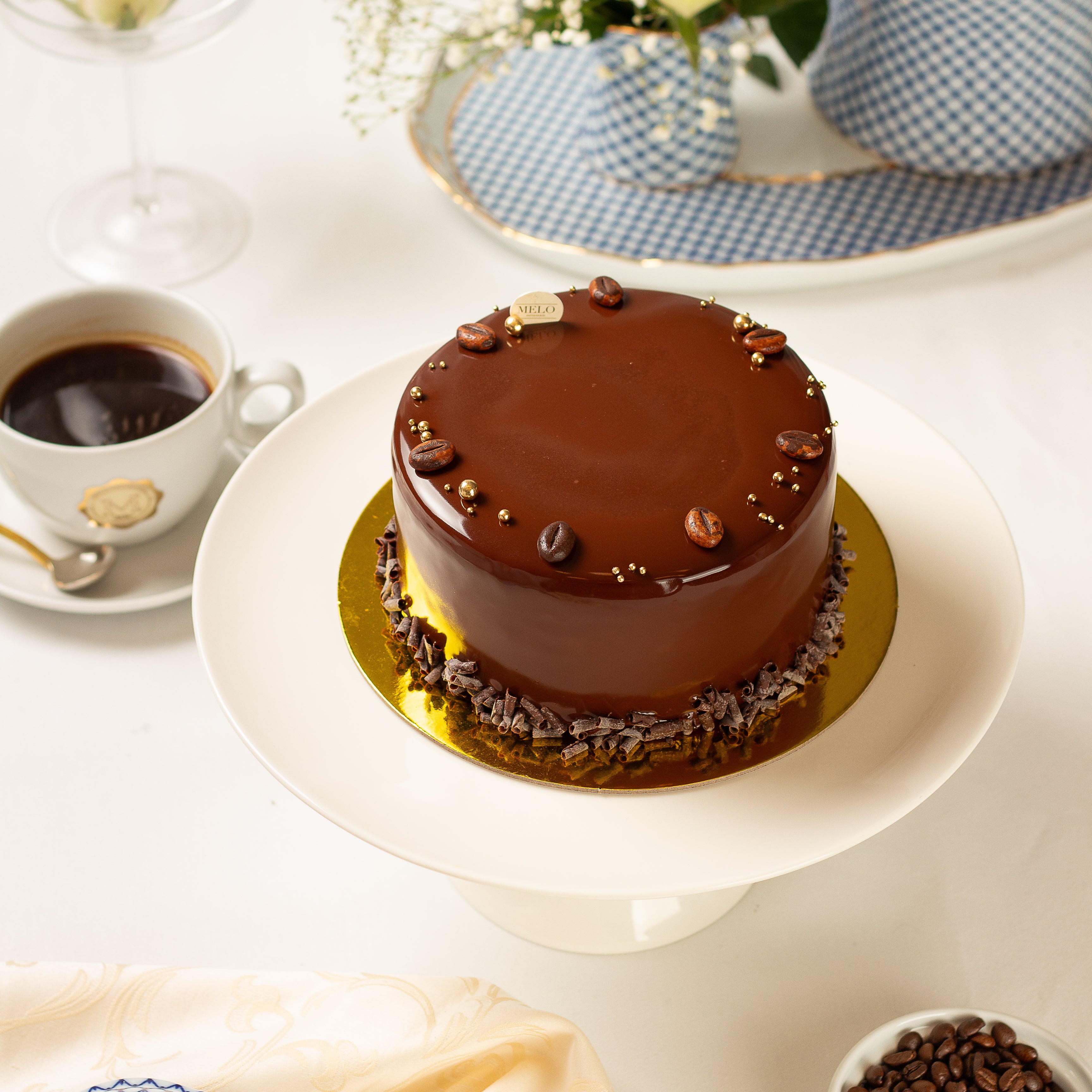 Chocolate cake on a white stand with a cup of coffee and a checkered dish in the background.