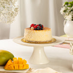 Decorative cake with berries on a white stand, surrounded by mango slices and flowers.