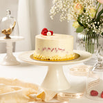 Small white cake with red berries on a white cake stand, surrounded by decorative items.
