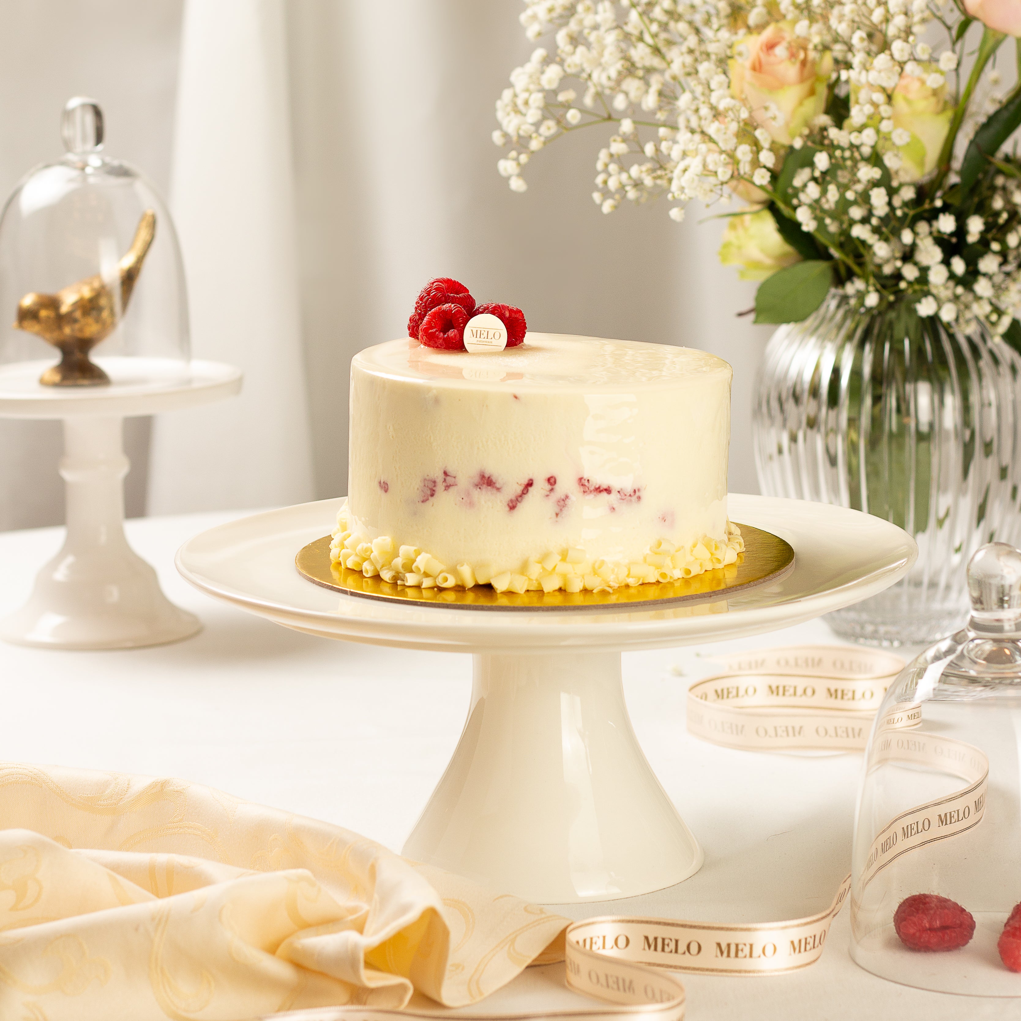 Small white cake with red berries on a white cake stand, surrounded by decorative items.