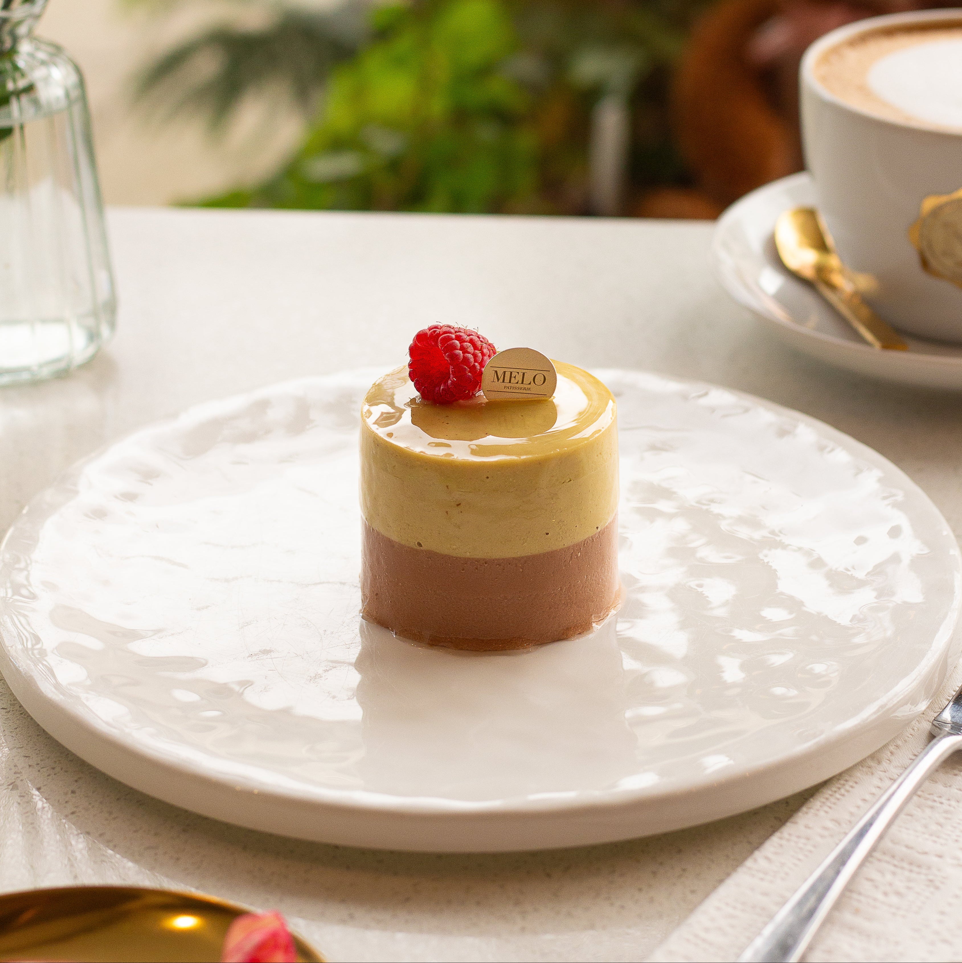 Small dessert with a raspberry on a white plate, accompanied by a cup of coffee and flowers in the background.