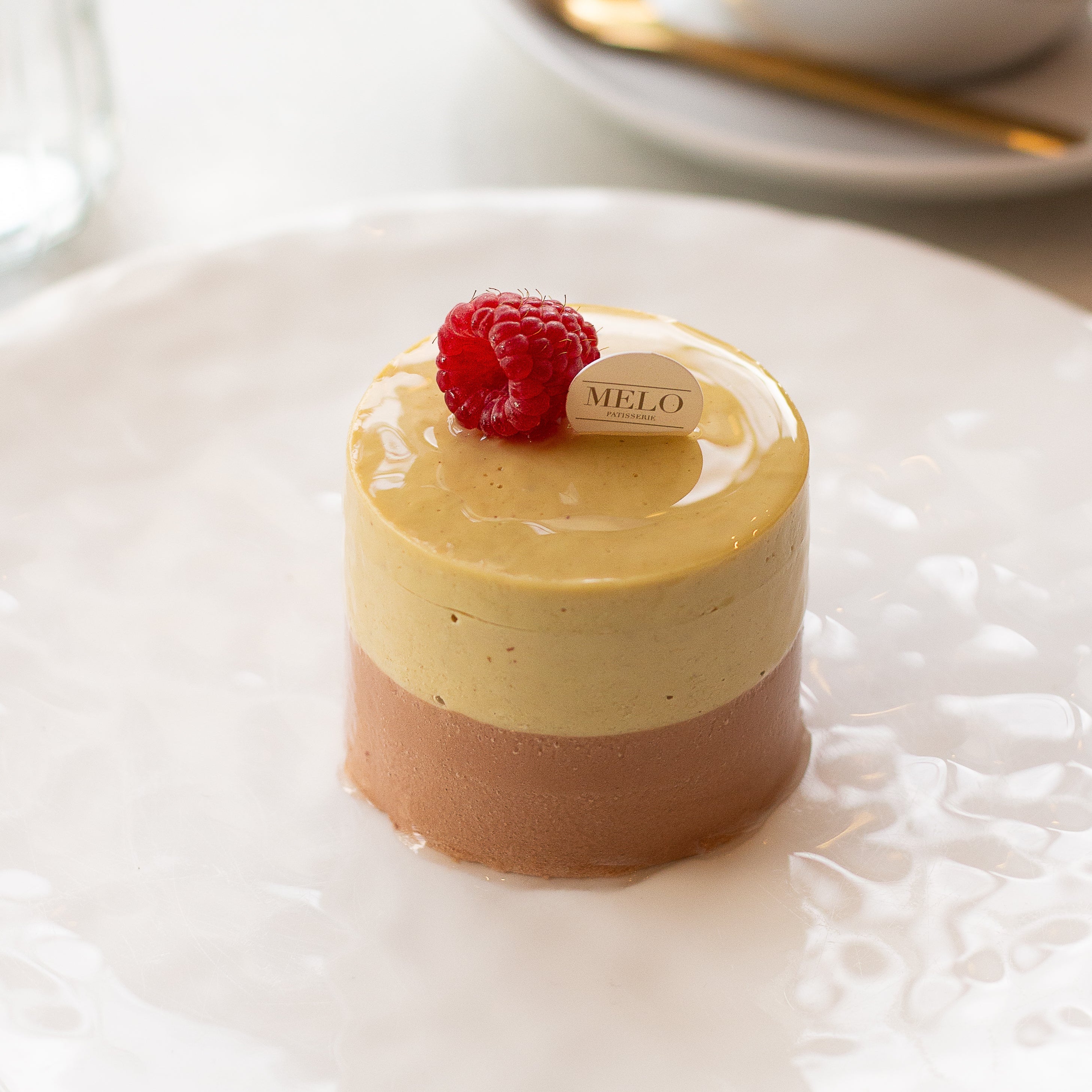 Two-layered dessert with a raspberry on top, served on a white plate with a cup of coffee in the background.
