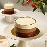 Triple layered chocolate cake on a white stand with flowers in the background