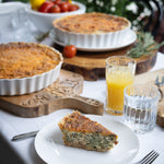 Quiche on a plate with glasses of juice and a bowl of tomatoes on a table outdoors.