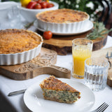 Quiche on a plate with glasses of juice and a bowl of tomatoes on a table outdoors.