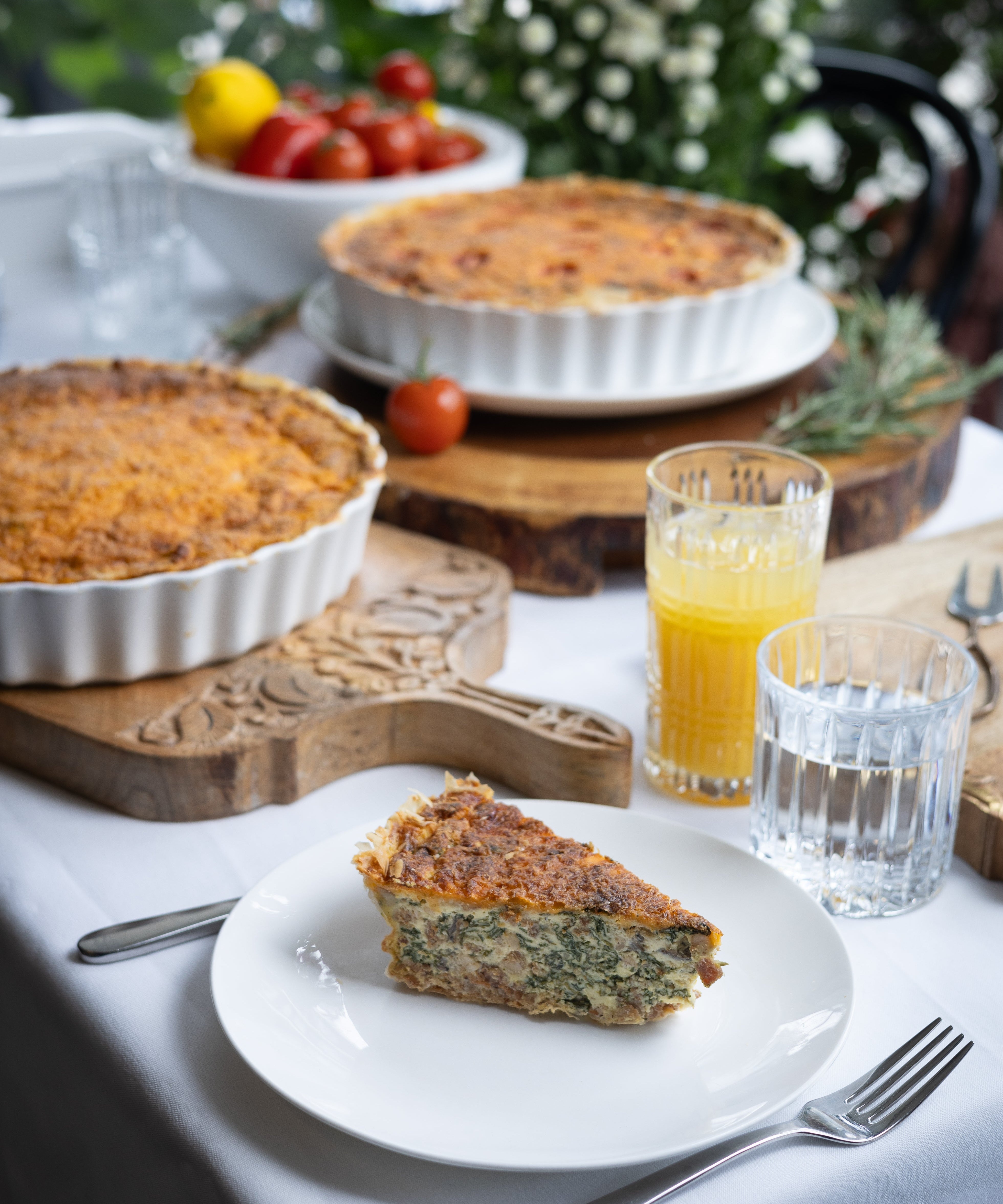 Quiche on a plate with glasses of juice and a bowl of tomatoes on a table outdoors.