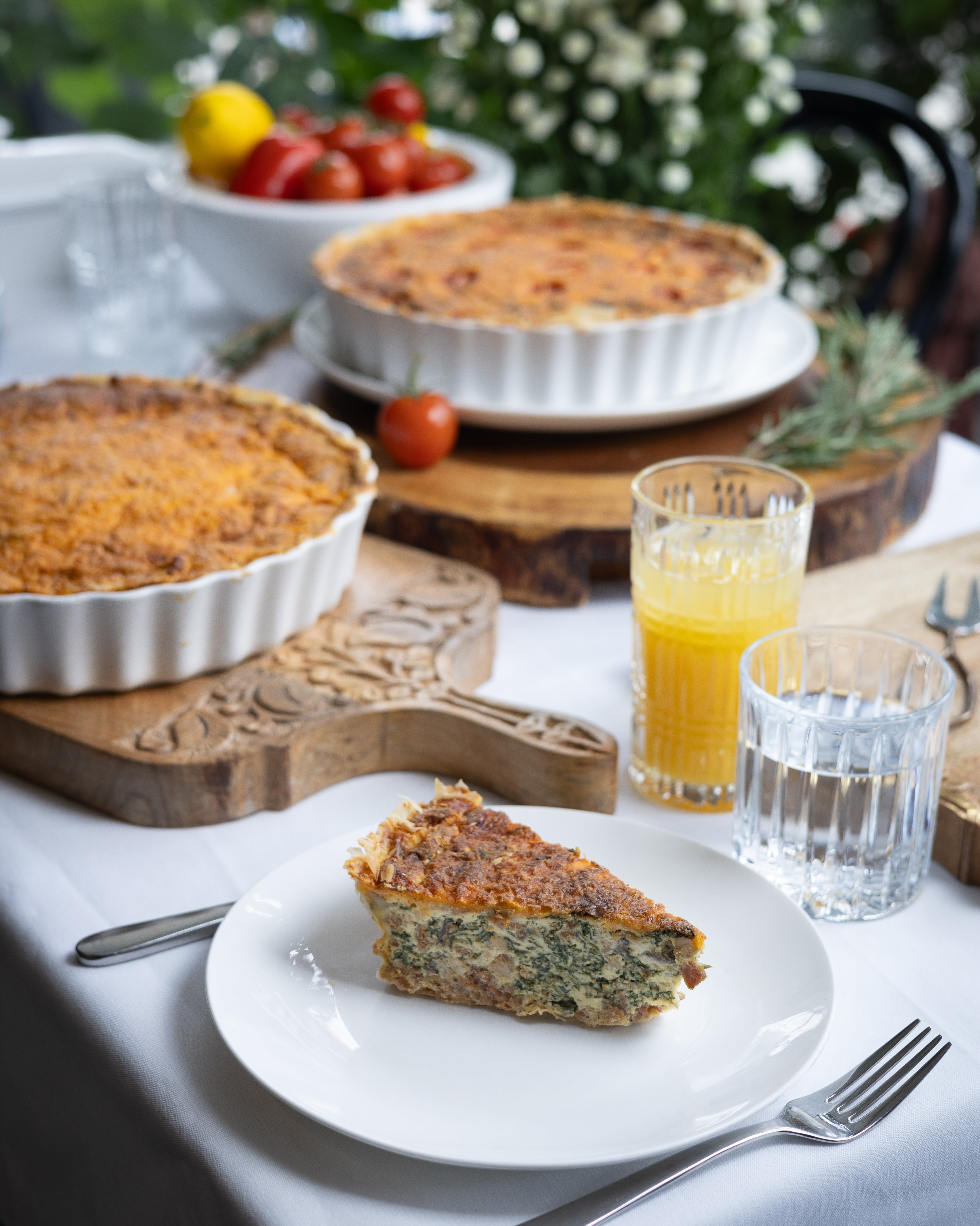 Quiche on a plate with glasses of juice and a bowl of tomatoes on a table outdoors.